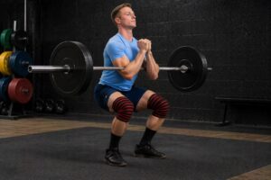 Powerlifter performing Zercher squat in a gym with barbell held in elbow crease, maintaining upright posture and control with knee wraps during mid-squat position