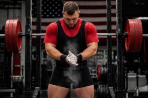 A powerlifter stands in front of a heavily loaded barbell in a squat rack, rubbing chalk onto their hands while focusing before an attempt. Wearing a lifting singlet, belt, and wrist wraps, they prepare mentally and physically for a heavy lift. An American flag hangs in the background, reinforcing the competitive strength-sport setting. The image represents performance readiness, concentration, and strong execution when handling maximal loads.
