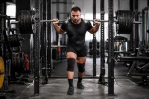 Heavy squat setup by competitive powerlifter with loaded barbell, lifting belt, and knee sleeves inside a gym power rack before unracking the weight.