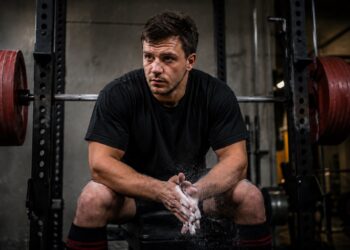A powerlifter sits on a bench inside a squat rack, chalking their hands and focusing before training. Heavy barbells loaded with red weight plates are positioned on either side, emphasizing a strength-training environment. The athlete's posture and concentration suggest preparation for upper-body training, illustrating the role of triceps work in building a stronger bench press.