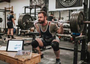 A focused powerlifter performing a heavy barbell back squat in a gritty gym, with a tablet displaying workout research data and training notebooks resting on a wooden bench nearby, bridging scientific data with practical strength training.